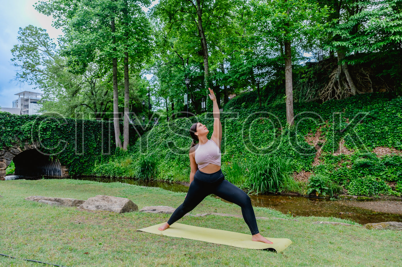 Young woman in yoga pose at a lush park, stone bridge background